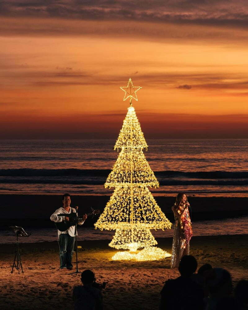 A special concert, set on the beach with a singer and musician playing acoustic guitar. Beautiful light-up Christmas tree on the sand and beautiful sunset light.